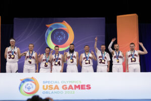 A basketball team is celebrating on a stage at the Special Olympics USA Games in Orlando 2022. The team members are standing in a line, wearing white uniforms with medals around their necks. They are raising their arms in victory, some giving a thumbs-up. Behind them is a backdrop featuring the Special Olympics logo and colorful design elements. The front of the stage has a sign that reads "Special Olympics USA Games Orlando 2022."